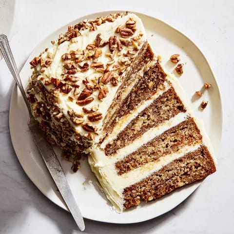 Golden, cream cheese-frosted layers of Hummingbird Cake studded with toasted pecans on a rustic table.