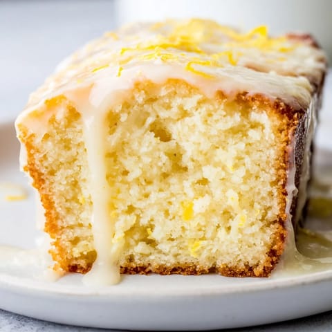 A moist slice of Lemon Pound Cake on a plate, crumbly texture revealed alongside a cup of tea for an afternoon treat.