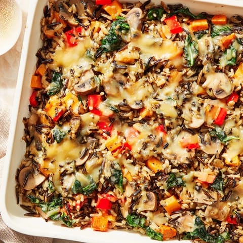 A spoon lifts a cheesy scoop of Wild Rice Casserole, revealing sautéed mushrooms, carrots, and red peppers.
